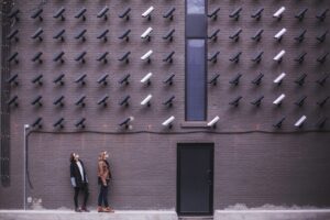 A vast array of surveillance cameras look down on two women who are looking up at them.